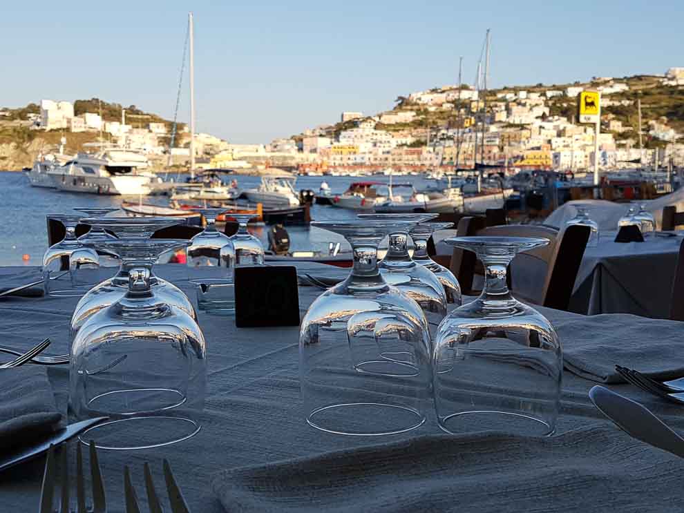 In primo piano, bicchieri sul tavolo di un ristorante di Ponza con vista sul porto.