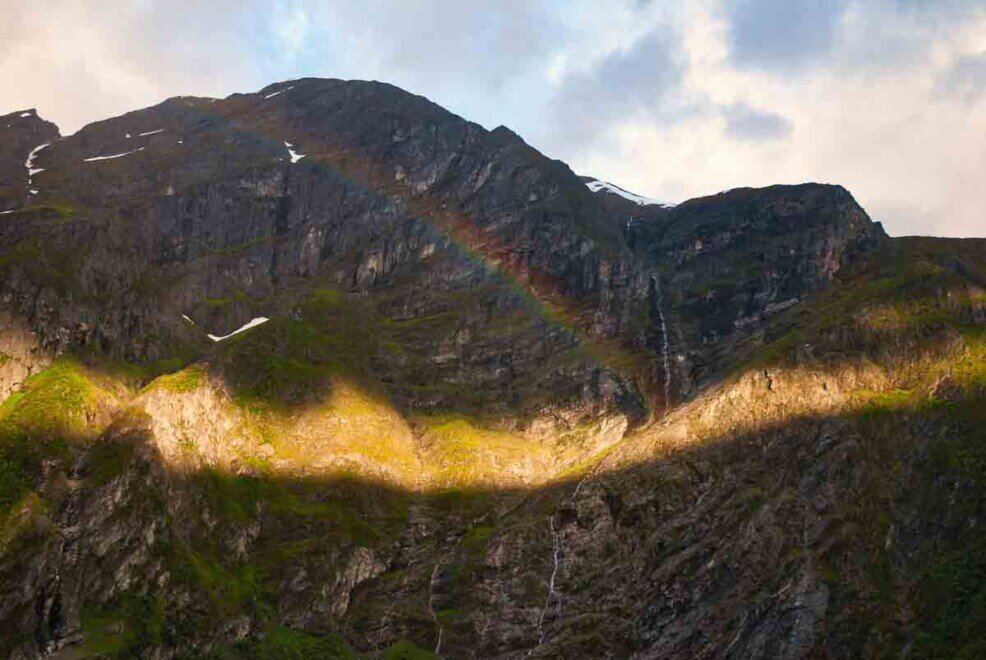 Geriangerfjiord-arcobaleno Geiranger Fjord: in uno spiraglio fra le nuvole filtra la luce del sole, e un arcobaleno si riflette sul costone della montagna.