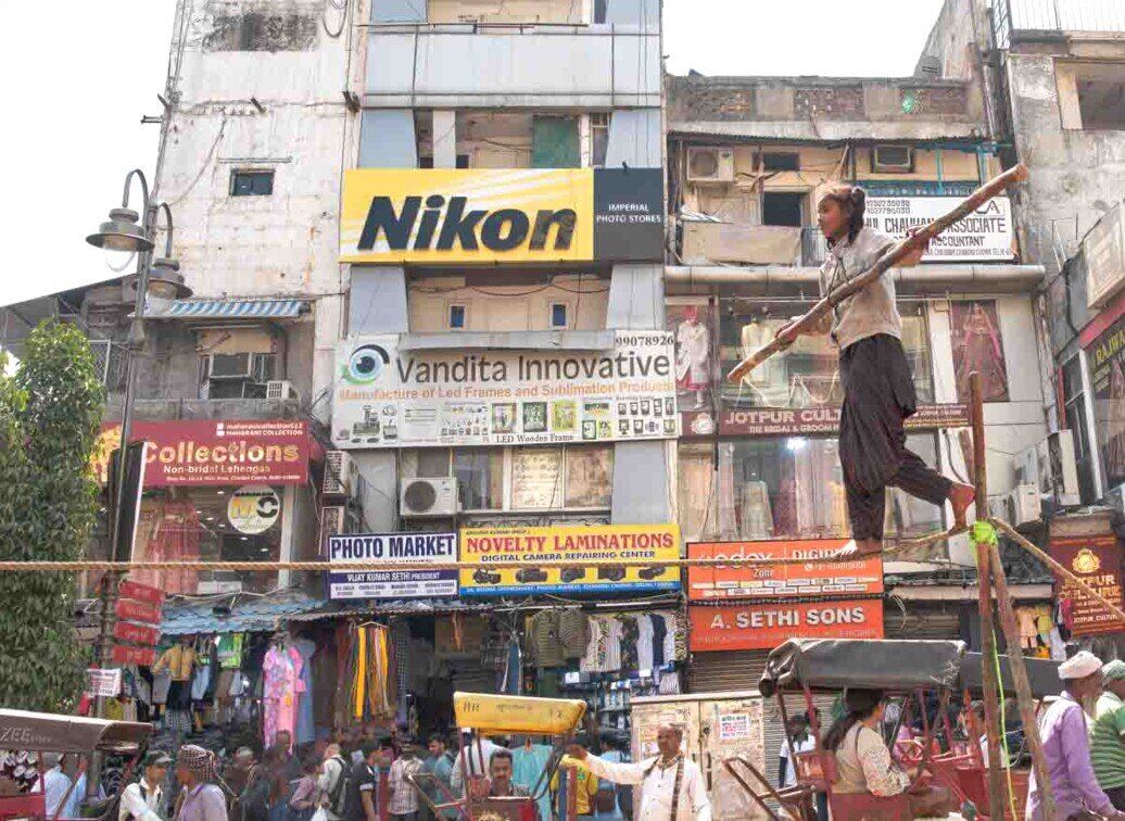 Ragazzina in equilibrio su una corda nella Delhi vecchia, con il vivace mercato di Chandni Chowk sullo sfondo.
