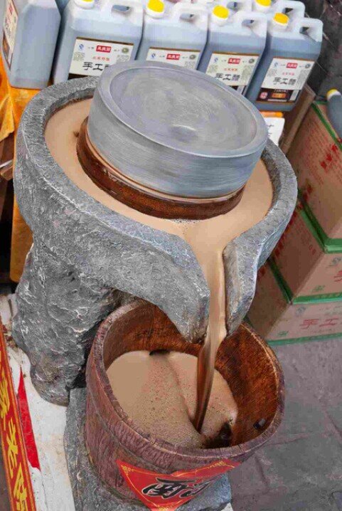 Fontana in pietra a Pingyao con liquido scuro che simula il processo tradizionale di produzione dell’aceto di Shanxi, con bottiglie in vendita sullo sfondo.
