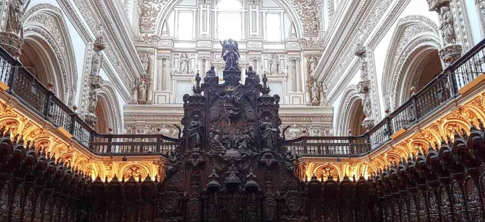 Platea del coro della Moschea-Cattedrale di Cordova con stalli in legno scuro finemente intagliati disposti su più livelli, decorazioni rinascimentali scolpite, grande leggio centrale e cattedra vescovile sul fondo, illuminata dalla luce naturale che filtra tra le alte volte della navata cristiana inserita nella struttura della moschea.