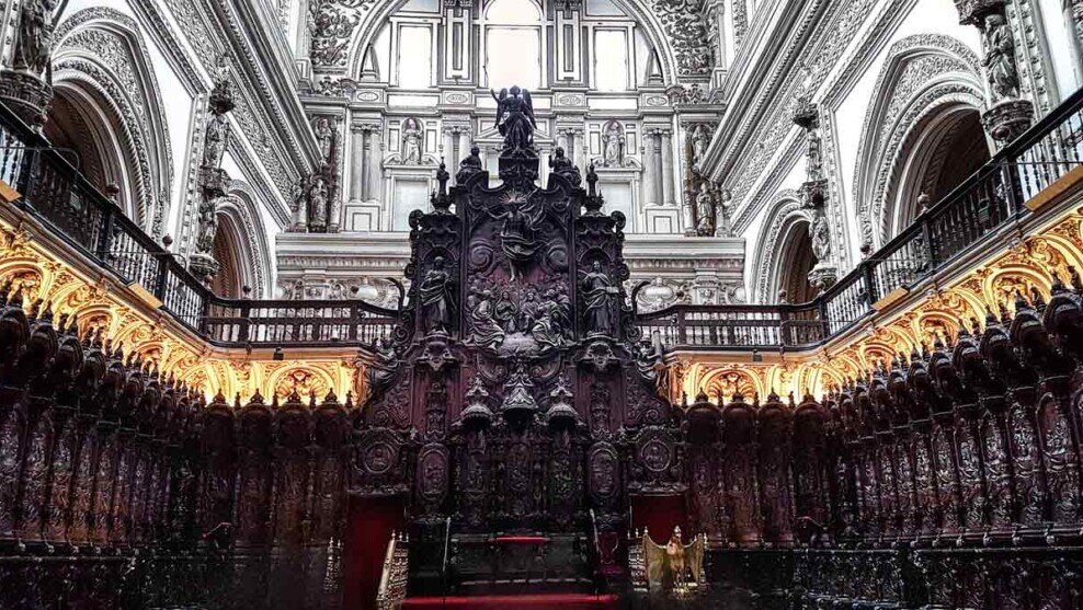 Platea del coro della Moschea-Cattedrale di Cordova con stalli in legno scuro finemente intagliati disposti su più livelli, decorazioni rinascimentali scolpite, grande leggio centrale e cattedra vescovile sul fondo, illuminata dalla luce naturale che filtra tra le alte volte della navata cristiana inserita nella struttura della moschea.