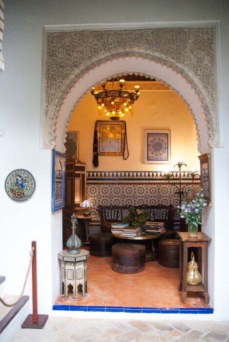 Un arco a ferro di cavallo, elemento tipico dell'architettura ispano-araba con le pareti decorate con piastrelle geometriche (zellige) e motivi in stucco intagliato. Lo spazio è arredato con divani bassi, tavolini intarsiati e illuminato da un lampadario in stile orientale.