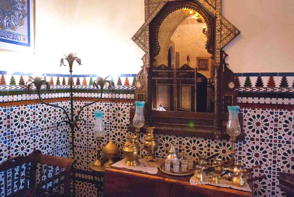L'ambiente è caratterizzato da pareti piastrellate con motivi geometrici (azulejos), un grande specchio in stile moresco e oggetti in ottone come teiere e vassoi.