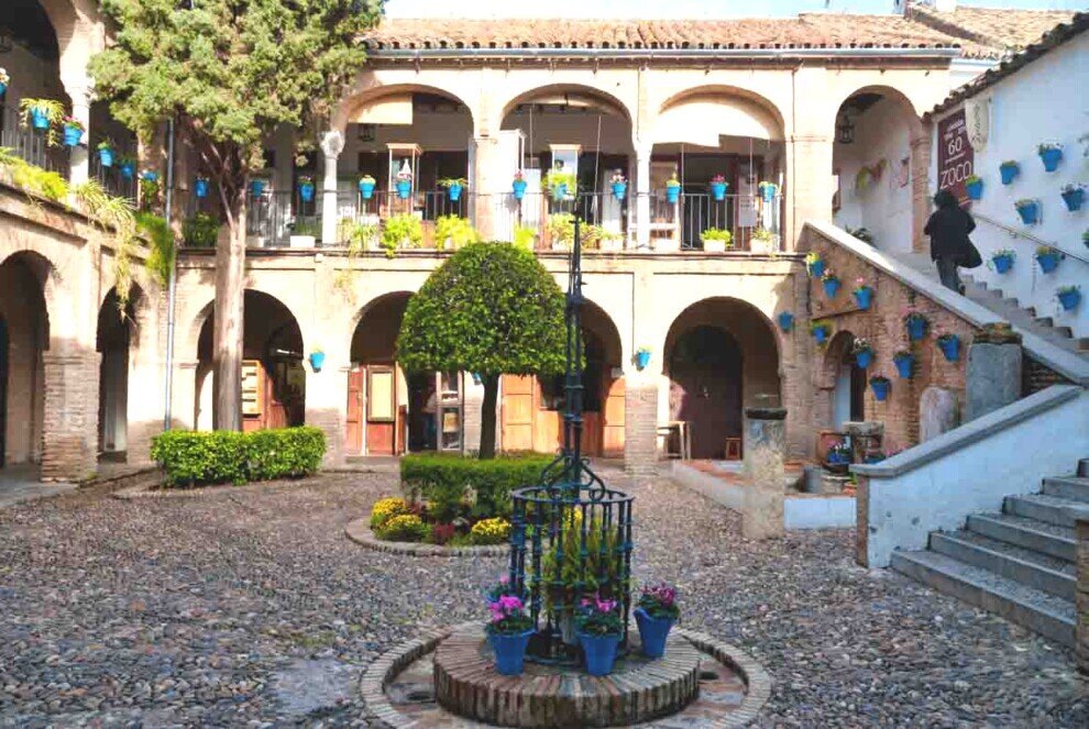 Cortile interno dello Zoco de la Judería a Córdoba con archi in pietra, vasi blu alle pareti, fontana centrale e pavimentazione in ciottoli.