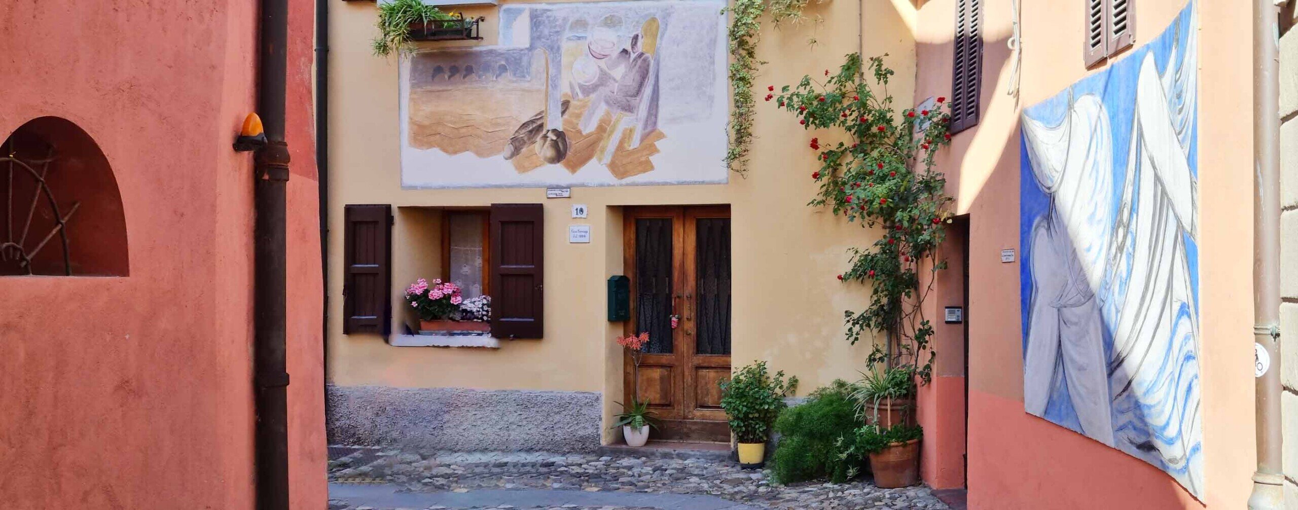 Una stretta via adornata da fiori alle porte e ai davanzali delle finestre con i bella mostra due murales dipinti sulle facciate.