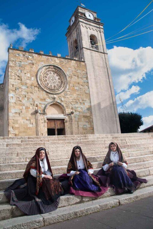 La facciata della chiesa di San Nicola con tre ragazze in costume sardo sedute sulla gradinata.