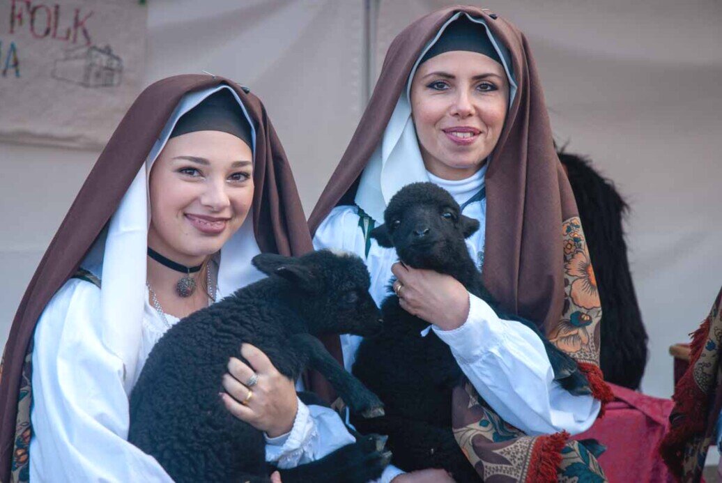 Due ragazze sorridenti in costume sardo con in braccio due piccoli agnellini neri.