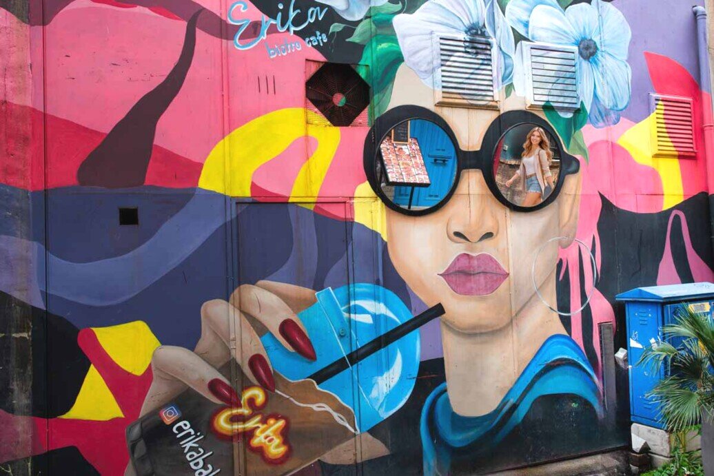 Murale che raffigura il volto di una ragazza con dei grandi occhiali che beve una bevanda da una cannuccia. Le lenti degli occhiali sono in realtà degli specchi che riflettono tutto ciò che si trova di fronte.