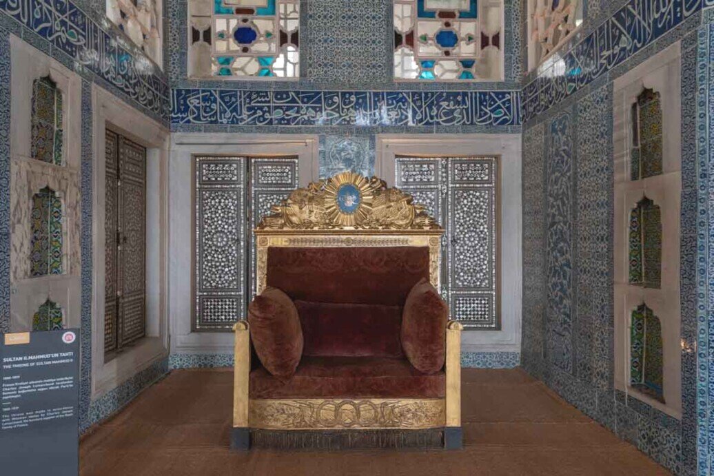 Una larga poltrona con bordi dorati e uno stemma, foderata in rosso con due cuscini ai lati dello stesso colore.