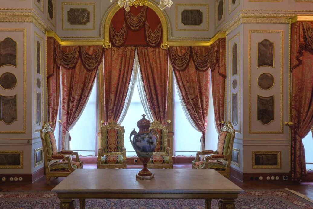 Un tavolo rettangolare in legno con sopra un vaso decorato, e sullo sfondo un finestrone a tutta altezza con tende rosse. Ai lati due poltrone dorate riccamente addobbate.
