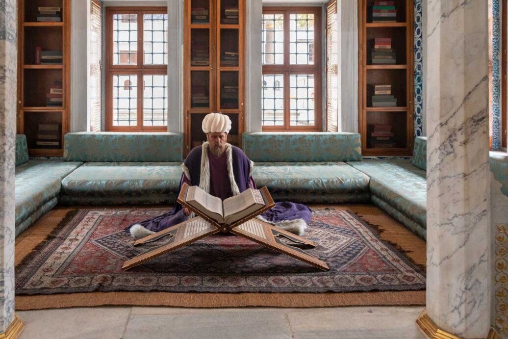 Un manichino con turbante seduto su un tappeto in una stanza circondata da libri.