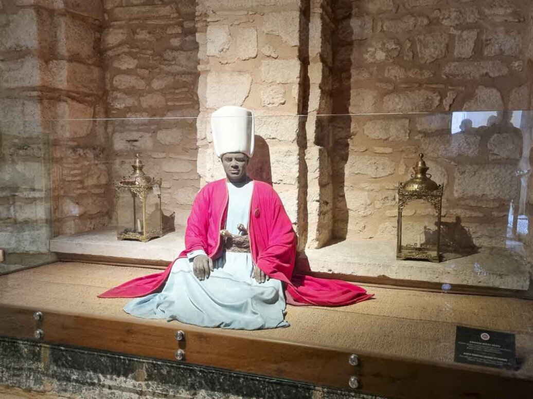 Un manichino vestito da eunuco seduto sul pavimento in legno del suo appartamento.