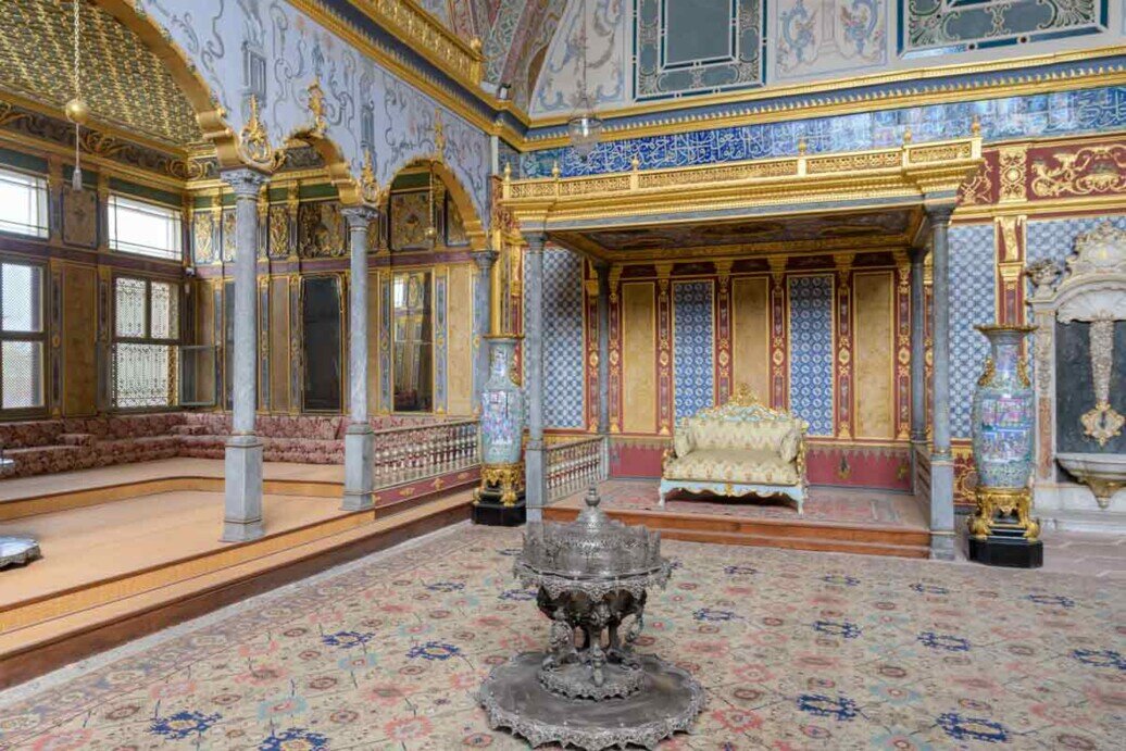 Interno della Sala delle Udienze del Palazzo Topkapi a Istanbul, con ricchi rivestimenti ottomani, pareti decorate con motivi geometrici e floreali, colonne finemente ornate e un divano posto al centro della stanza.