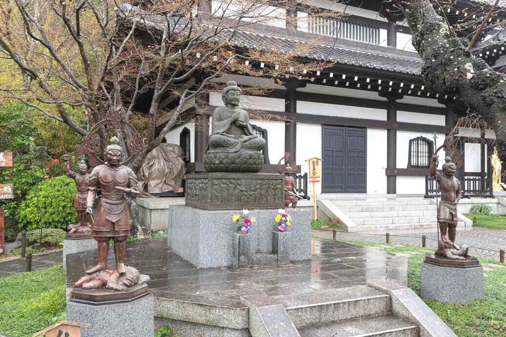 Tempio giapponese con statua del Buddha seduto su piedistallo in pietra, circondato da statue di guardiani, alberi spogli e un edificio tradizionale sullo sfondo; il terreno è bagnato dalla pioggia.