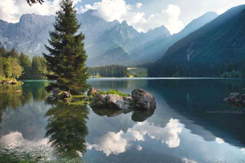 Lago di Fusine su cui si specchia il paesaggio Un lago su cui si specchiano le nuvole e un albero con sullo sfondo alte montagne.