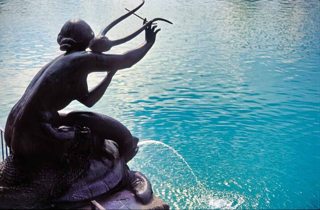 Silhouette della figura della statua in bronzo raffigurante una musicista (sirena) su una tartaruga contro l'acqua turchese, mettendo in risalto i dettagli della scultura e del getto d'acqua.