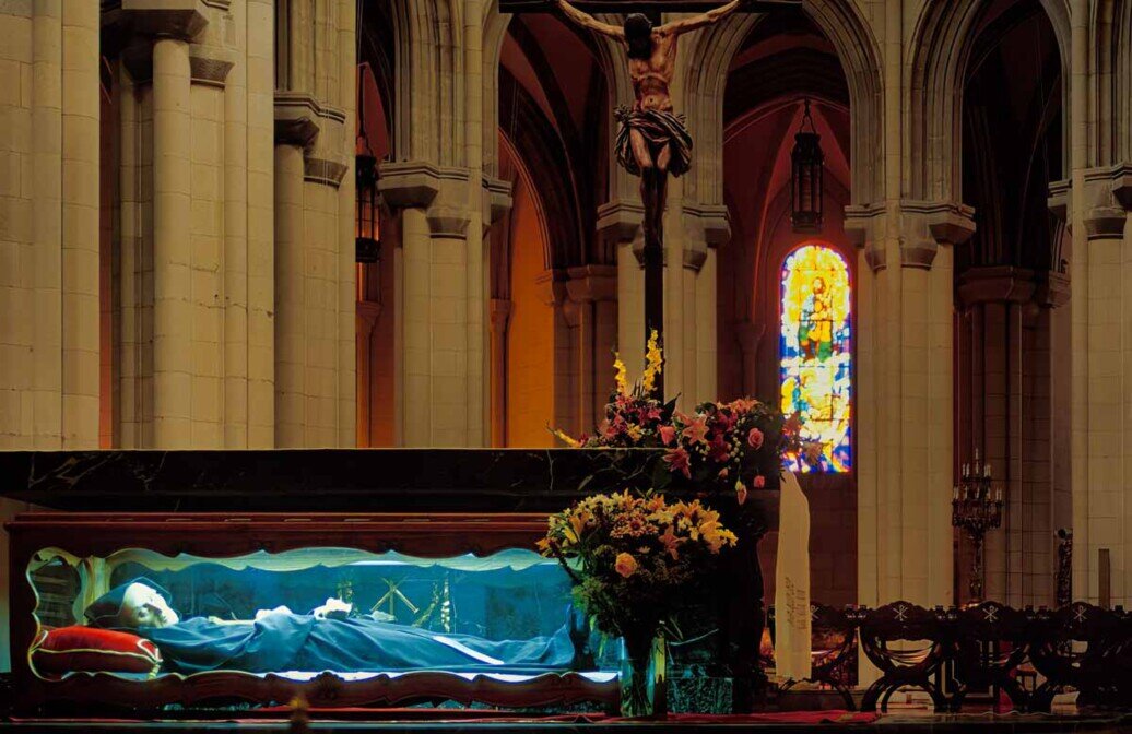 Interno della Cattedrale dell’Almudena a Madrid, tra colonne e volte, con la statua del corpo della Madonna sdraiata supina in primo iano dentro una teca illuminata.