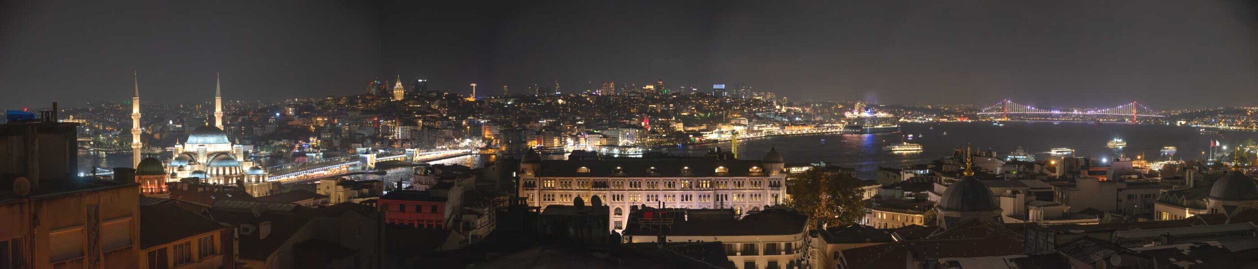 Panorama notturno su Istambul affacciato sul bosforo, con sullo sfondo una moschea illuminata un ponte, un palazzo, e un altro pronte ad una campata sullo sfondo che unisce la Istambul europea da quella asiatica.