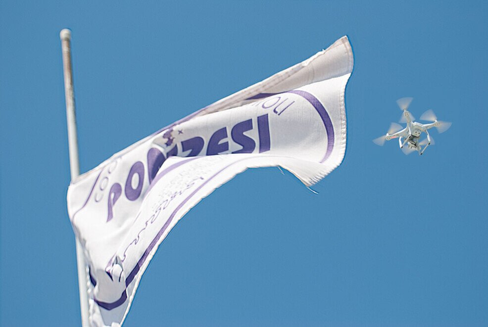 Bandiera della cooperativa pescatori ponzesi che si staglia sul cielo, a fianco di un drone che sorvola e che riprende la scena della processione in onore di San Silverio a Ponza.