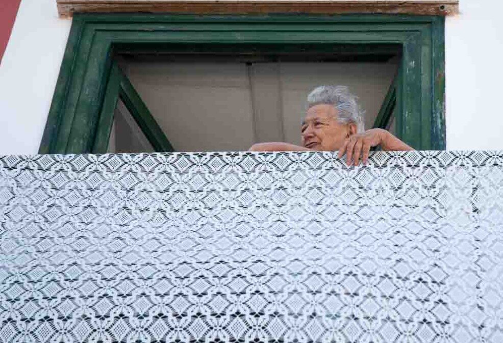 Una signora anziana seduta al balcone per assistere alla processione di San Silverio a Ponza ha appeso una coperta ricamata in onore del santo.