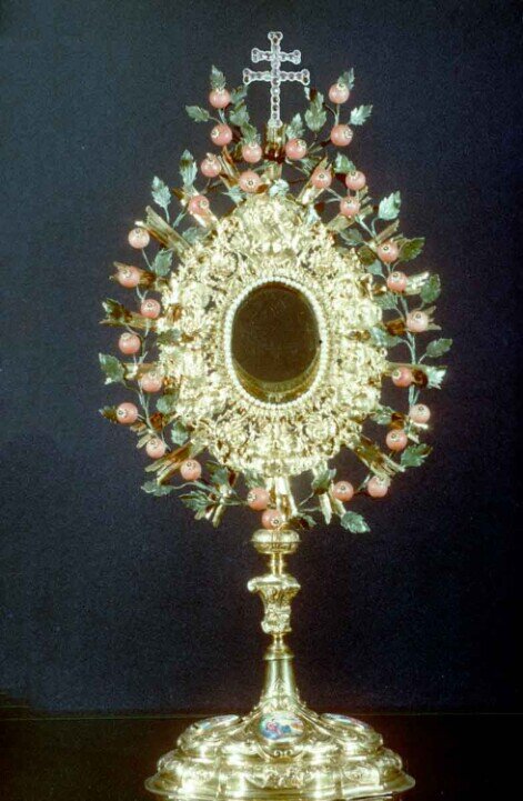Ostensorio in argento dorato con ricche decorazioni a rilievo e piccoli smalti figurativi sulla base. La corona è ornata con rami di foglie verdi e piccoli frutti rosati (simili a bacche o piccoli melograni).