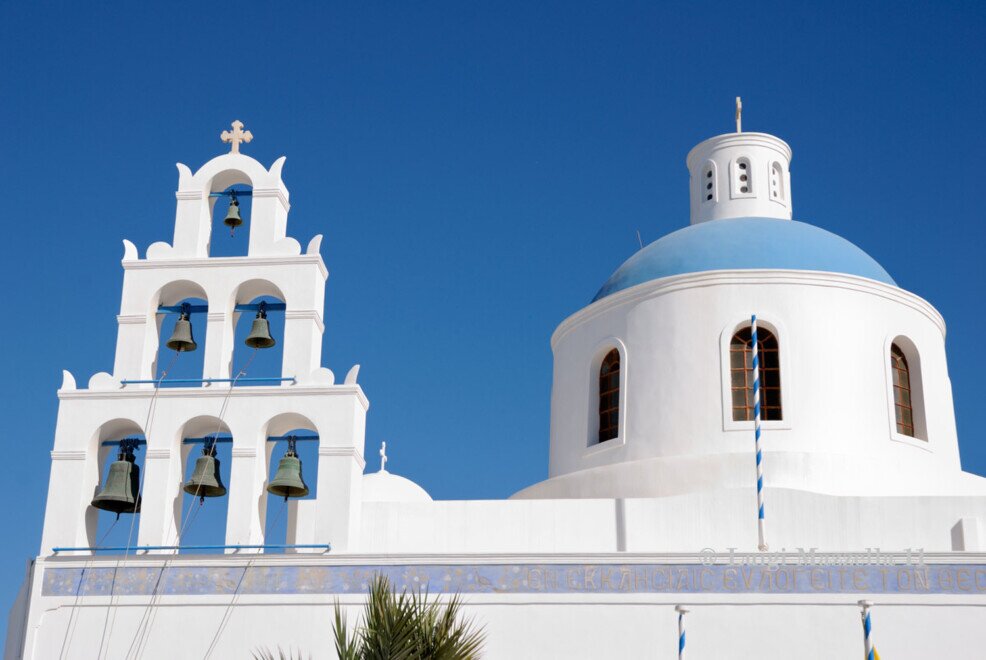 Chiesa bianca con cupola blu e campanile a più archi con campane, tipica architettura di Santorini sotto cielo azzurro.