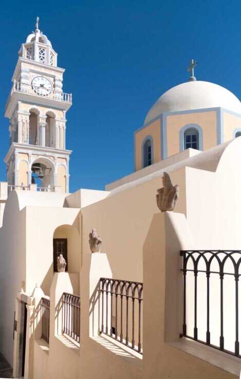 Cattedrale cattolica a Firà nell'isola di Santorini. Ha un campanile con orologio e a fianco la cupola della chiesa tutto di colore pastello con alcuni bordi azzurri.