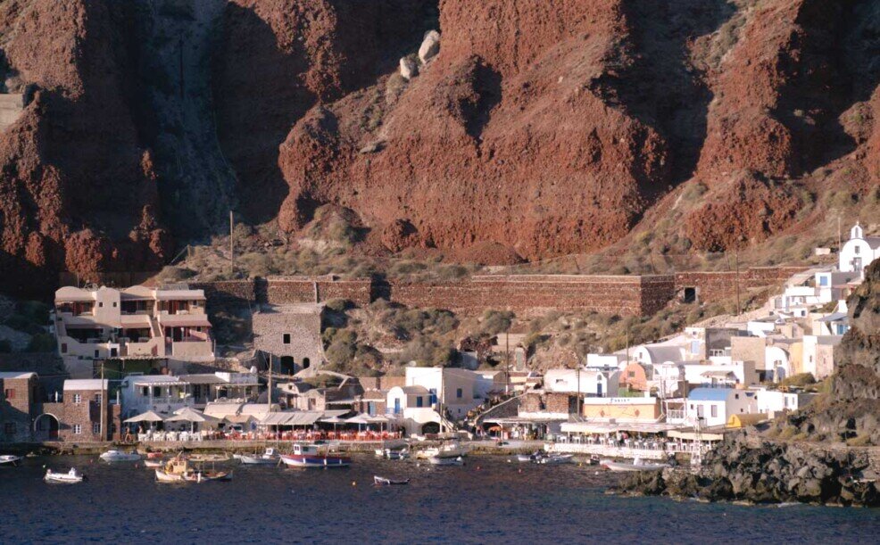 La baia di Amoudi a Santorini dove c'è un porticciolo e delle case e da dove si innalzano alte pareti di colore rossastro testimonianza di una grande eruzione vulcanica del passato.