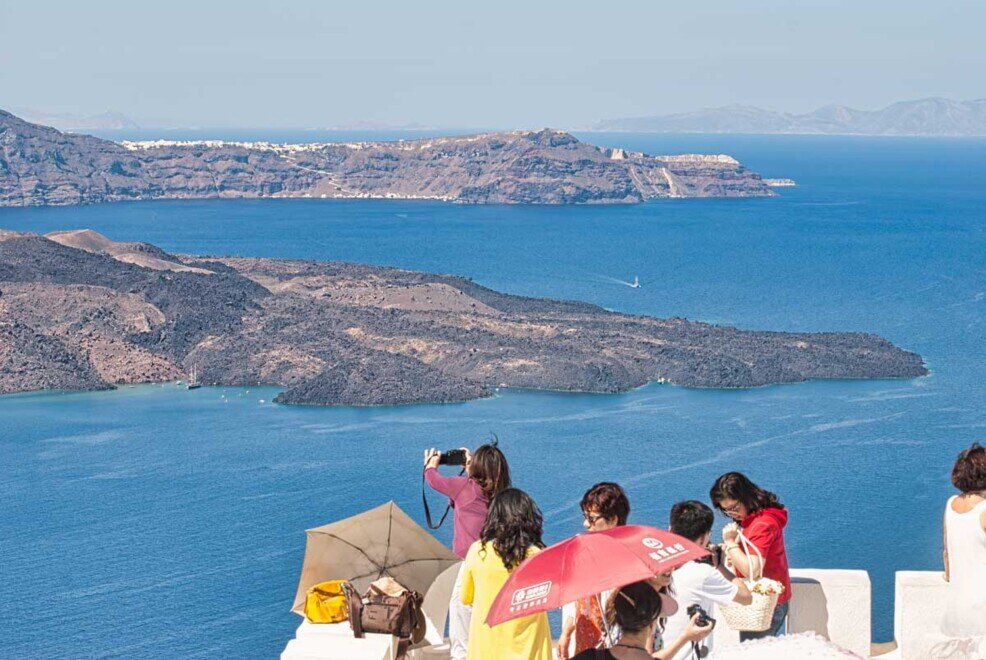 Santorini-un gruppo di turisti fotografano dall'alto di uno spiazzo il panorama sulle scogliere della caldera.