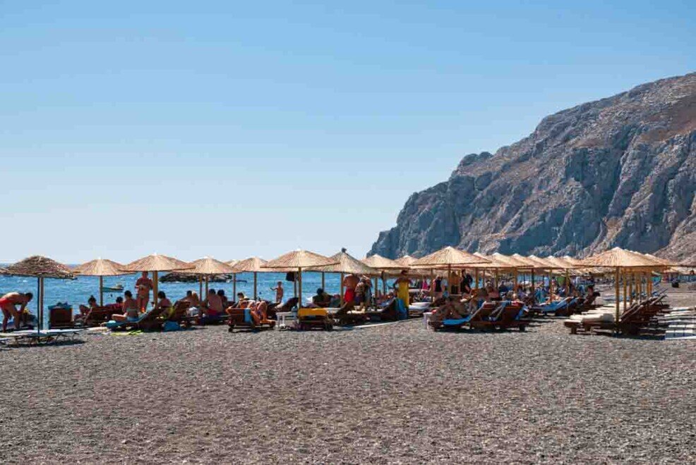 Ombrelloni gialli piantati sulla sabbia nera di origine vulcanica, con bagnanti, nella spiaggia di Kamari a Santorini.