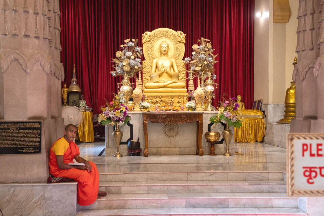 La statua dorata del Buddha al tempio di Sarnath Un monaco seduto con le mani unite davanti a un Buddha dorato su una piattaforma di marmo.<br />