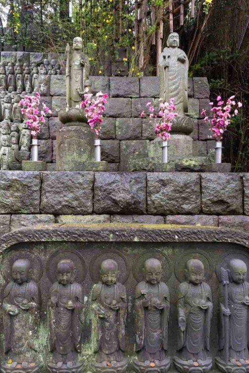 Statue di Jizō in pietra disposte su una parete a gradoni, decorate con fiori rosa, in un giardino del tempio giapponese.