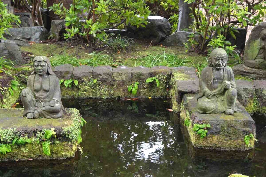 Due statue in pietra raffiguranti figure sedute ai lati di una piccola vasca d’acqua, in un giardino verde con muschio e piante.